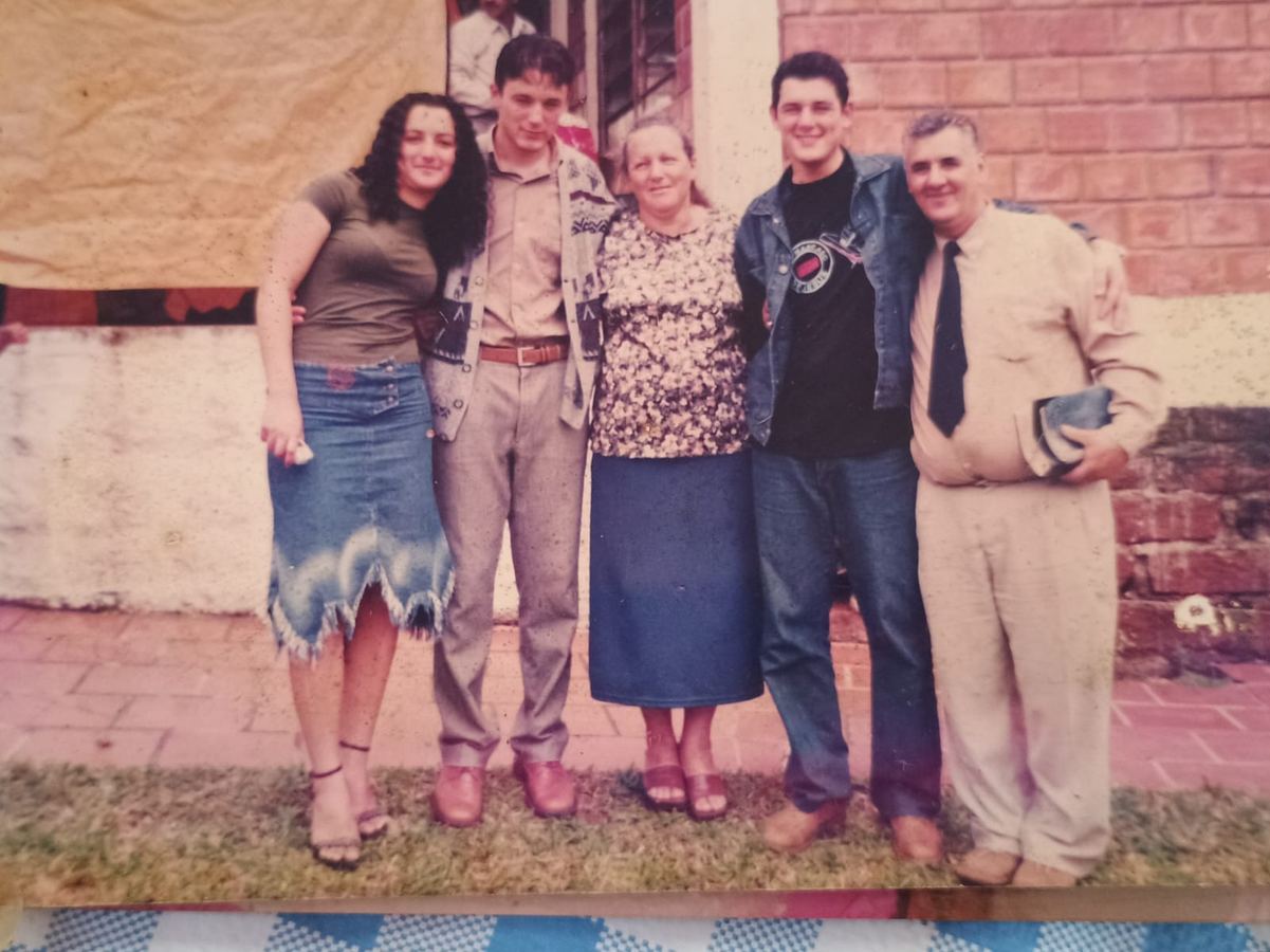 Fotografia de família em pé diante de uma casa de tijolos, com jovens e adultos abraçados