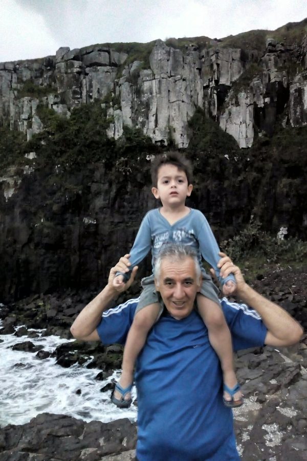 Um senhor com uma criança nos ombros diante de rochas e ondas