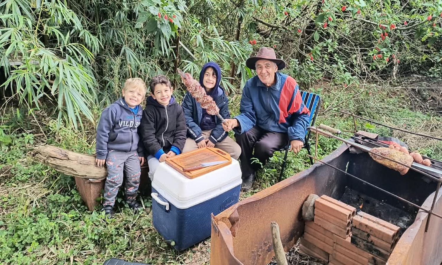 Crianças e um senhor de chapéu ao lado de uma caixa térmica, em área rural