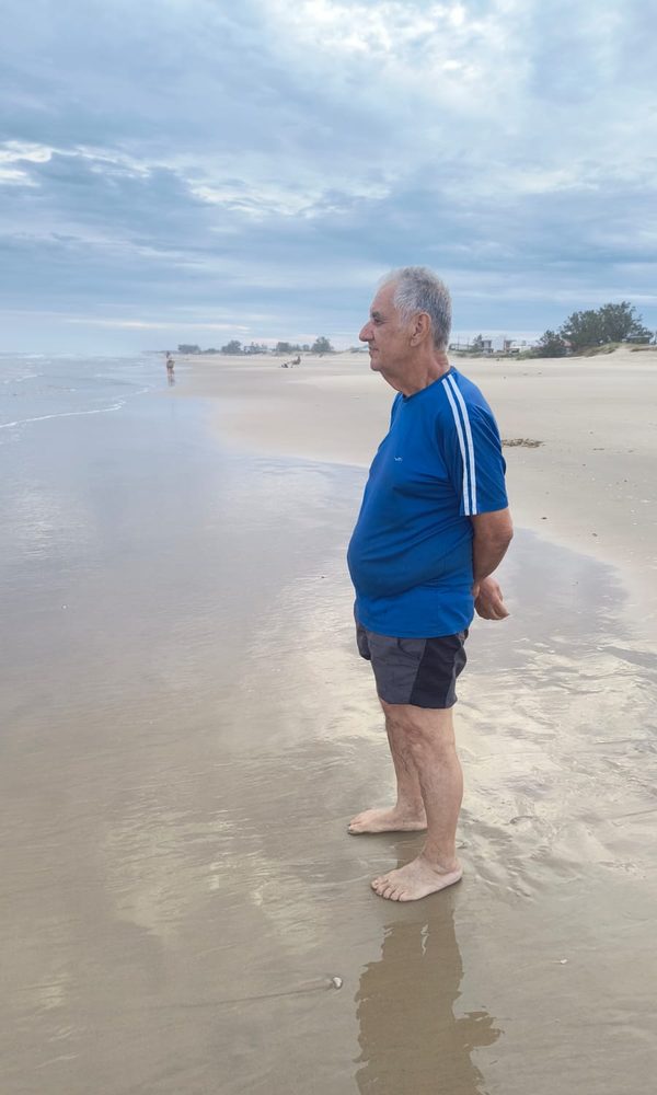Sinval de perfil, de pé na beira da praia, olhando o mar