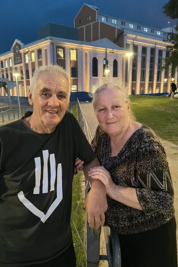 Sinval e uma mulher abraçados à noite, em frente a um prédio iluminado